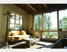 Wohnraum mit rustikalem Holzinterieur, großen Fenstern mit Blick auf eine bewaldete Landschaft und ein Gewässer. Im Vordergrund stehen ein gelbes Sofa, ein niedriger Holztisch und eine Stehlampe, mit Gitarren und gerahmten Bildern an der Wand angelehnt.