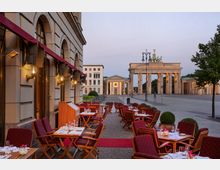 Außenbereich eines Restaurants mit gedeckten Tischen und roten Stühlen entlang einer steinernen Fassade, im Hintergrund das beleuchtete Brandenburger Tor in Berlin. Die Szene zeigt eine ruhige Abendstimmung mit wenigen Personen und Bäumen in der Umgebung.