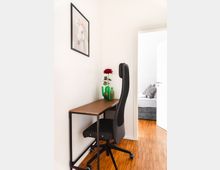 Schreibtischecke in einem hellen Raum mit einem minimalistischen Holztisch, einem schwarzen Bürostuhl und dekorativen Vasen sowie einer roten Blume. An der Wand hängt ein Bild eines weißen Pferdes, im Hintergrund ist eine offene Tür zu einem Schlafzimmer mit einem Bett sichtbar.