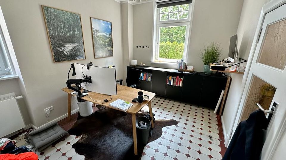 Arbeitszimmer mit einem Holztisch in der Mitte, zwei Monitoren und Kabelmanagement-Armen. Der Raum hat cremefarbene Wände, ein großes Fenster mit Blick auf grüne Bäume, eine niedrige schwarze Schrankwand mit Büchern und Dekoration, sowie einen rot-weißen Fliesenboden und eine helle Deckenleuchte.