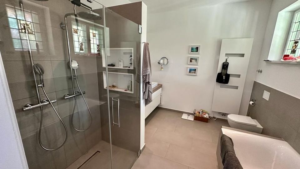 Modernes Badezimmer mit einer großen, verglasten Doppeldusche, einer Badewanne, einem Wand-WC und einem weißen Waschtisch. Die Wände sind weiß, Böden und Duschwände sind mit großen, grauen Fliesen verkleidet; kleine Fenster mit farbigem Glas lassen Tageslicht herein.
