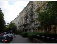 Straßenansicht mit einer Reihe von mehrgeschossigen Altbauhäusern, die verzierte Fassaden und Balkone haben, teils mit Blumenschmuck. Rechts säumen Bäume und Hecken den Gehweg, während links geparkte Autos entlang der Straße stehen.