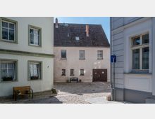 Eine kleine gepflasterte Gasse mit Wohnhäusern im Blickfeld. Links ein hellgrünes Gebäude mit Blumenkästen an den Fenstern und einer Holzbank davor, im Hintergrund ein cremefarbenes Haus mit braunem Garagentor.