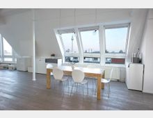 Ein heller, moderner Dachgeschossraum mit großen schrägen Fenstern, die eine Stadtansicht zeigen, inklusive eines markanten Fernsehturms. Im Vordergrund steht ein Holztisch mit vier weißen Stühlen, umgeben von minimalistischen weißen Möbeln.