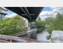 Eine Brücke mit sichtbarer Unterkonstruktion, die über einen schmalen Kanal mit befestigten Ufern und Graffiti führt. Im Hintergrund sind Bäume, ein Gebäude des Deutschen Technikmuseums sowie andere moderne Gebäude zu sehen.
