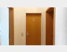 Ein Flur mit drei Türen aus hellem Holz. Die mittlere Tür hat ein Schild mit einem Rauchverbot-Symbol, die Wände sind weiß gestrichen.