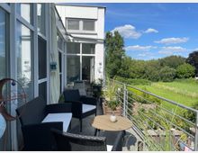 Ein moderner Balkon mit Glasgeländern und Sitzmöbeln aus Rattan, umgeben von Pflanzen in Töpfen. Im Hintergrund sind eine grüne Wiese, Bäume und ein blauer Himmel mit wenigen Wolken zu sehen.