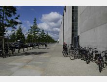 Breiter Gehweg mit Betonplatten und Kieselsteinpflaster, gesäumt von einer hohen Betonmauer rechts und einer Reihe junger Bäume links entlang parkender Autos. Fahrräder sind an Ständern entlang der Mauer abgestellt, und eine Person geht in der Entfernung.