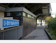 Außenansicht des U-Bahnhofs Hallesches Tor in Berlin mit einem blauen Stationsschild im Vordergrund. Das Bauwerk zeigt eine überdachte Bahnsteigstruktur mit Metallträgern und umliegender Vegetation.