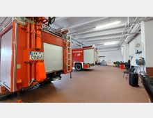 Innenansicht einer Feuerwehrgarage mit zwei geparkten roten Feuerwehrfahrzeugen. Der Raum hat eine weiße Decke mit Beleuchtung, helle Wände, einen braunen Boden und an den Seiten Lagergegenstände wie Schläuche, Stühle und Werkzeuge.