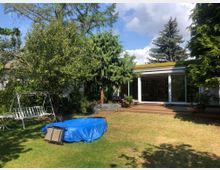 Ein Garten mit Rasenfläche, Bäumen und einer weißen Schaukelbank. Im Hintergrund befindet sich ein modernes Haus mit einer breiten Fensterfront, dahinter sind Bücherregale zu sehen.
