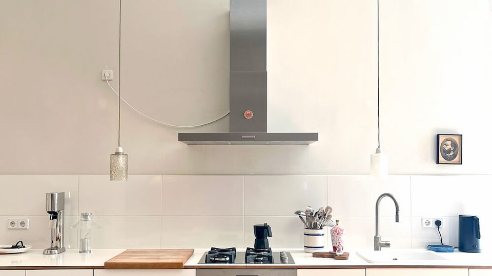 Minimalist white kitchen with a long counter and flat-front cabinets, featuring a built-in oven and gas cooktop beneath a stainless-steel range hood. Two pendant lights hang from the high ceiling, and a sink, small appliances, and utensils sit along the tiled backsplash.