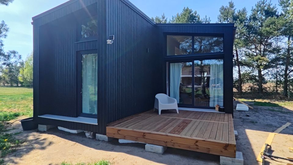 Modernes, schwarzes Holzhaus mit Satteldach und großflächigen Fensterfronten, davor eine erhöhte Holzterrasse auf Betonblöcken. Das Gebäude steht auf sandigem, teils bewachsenem Gelände am Rand einer Baumreihe unter blauem Himmel.