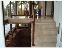 Treppenaufgang mit beigefarbenen Marmorstufen in einem Eingangsbereich. Rechts ein hölzernes Geländer, links ein Fensterbereich mit Ziegelmauerwerk und Topfpflanzen; im oberen Bereich Zierpflanzen und Dekorationen auf einem hellen Boden.