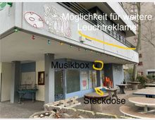 Gebäudeeingang mit überdachtem Vorbereich, Betonpfeilern und blauer Wand; darüber Balkone mit schwarzem Geländer. Im Vordergrund stehen mehrere runde Steintische und Stühle. An der Decke hängen bunte Glühbirnen, und an der Wand befindet sich ein leuchtender Schriftzug und Graffiti.