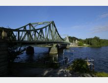Grüne Stahlbrücke (Glienicker Brücke) über einen Fluss, umgeben von Bäumen und Wiesen; im Vordergrund ein Bootsanleger mit einem Schild „Station Glienicker Brücke 1“.