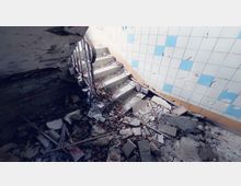 Zerfallenes Treppenhaus mit beschädigten Betontreppen und einem rostigen Metallgeländer. Die Umgebung ist von Schutt bedeckt, mit einer gekachelten Wand im Hintergrund, die blau-weiße Fliesen zeigt.