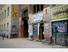 Straßenansicht eines heruntergekommenen Gebäudes mit Graffiti an den Wänden und Fenstern; ein gelbes Schild mit der Aufschrift „Auto Hof“ befindet sich an der Ecke. Der Eingangsbereich zeigt freiliegendes Mauerwerk und herumliegende Betonplatten.