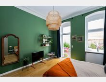 Schlafzimmer mit einer dunkelgrünen Wand, einem großen Spiegel, Holzfußboden und einem Bett mit orangener Decke. An der Wand hängen Regalboards mit Pflanzen und Deko, ein kleiner Fernseher steht auf einem Gestell, und durch bodentiefe Fenster fällt Blick auf eine Terrasse mit Pflanzen und Sitzmöbeln.