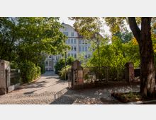 Eingang zu einem Schulgebäude mit einer breiten Torzufahrt aus Kopfsteinpflaster, flankiert von einem metallenen Zaun und von Bäumen gesäumt. Im Hintergrund ist ein großes, mehrstöckiges Gebäude mit weißen Fassaden und roten Dächern sichtbar, umgeben von dichtem Grün.