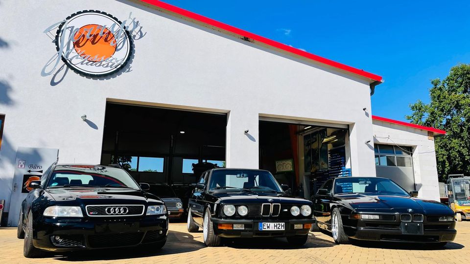 Frontansicht einer Autowerkstatt mit weißer Fassade und rotem Dachrand, zwei großen offenen Garagentoren und einem runden Logo über dem Eingang. Davor liegt ein gepflasterter Hof mit mehreren geparkten Autos; im Hintergrund sind blauer Himmel und Bäume zu sehen.