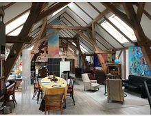 Ein großzügiger, offener Wohnraum in einem Dachgeschoss mit sichtbaren Holzbalken und schrägen Decken. Der Raum ist möbliert mit einem Esstisch, Sesseln, Sofas und dekorativen Elementen wie Gemälden und Regalen; große Dachfenster lassen Tageslicht herein.