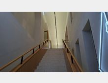 Blick von unten auf eine breite Innentreppe mit hellen Stein-Stufen und Holzgeländern, die zu einem schmalen, hohen Flur führt. Weiße Wände mit Nischen und Bögen sowie eine lange Deckenleuchte prägen den modernen Innenraum.