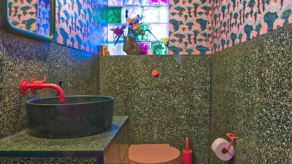 Small modern bathroom with terrazzo wall and floor tiles, a round black vessel sink with a bright red faucet, and a wall-mounted pink toilet. Patterned wallpaper and a colorful glass-block window provide vivid accents, with a mirror and globe light fixture above the vanity.