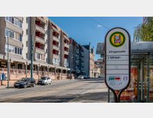 Straßenszene mit einer Straßenbahnhaltestelle „Klingestraße“ im Vordergrund, mehrstöckigen Wohngebäuden links und einer Straße mit geparkten Autos und Fahrspuren rechts. Im Hintergrund sind ältere Fassaden und ein Kirchturm sichtbar.