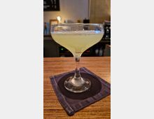 Ein Cocktailglas mit hellgrünem Getränk steht auf einem hölzernen Tresen, platziert auf einer dunklen Stoffserviette; im Hintergrund ist eine leicht unscharfe Bar mit Regalen und Beleuchtung erkennbar.