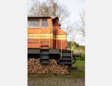 Seitenansicht einer alten, orangefarbenen Lokomotive mit gelben Streifen, die teilweise rostig ist. Darunter ist ein Stapel geschnittenes Brennholz zu sehen, im Hintergrund stehen Bäume mit wenigen Blättern.