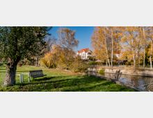 Ein Park mit einer Wiese, herbstlich gefärbten Bäumen, einer Holzbank und einem Fluss mit einer Steinmauer am Ufer. Im Hintergrund sind Häuser mit roten Ziegeldächern zu sehen.