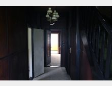 Dunkler Flur in einem Gebäude mit Holzverkleidung an den Wänden, einer Treppe mit dunklem Holzgeländer auf der rechten Seite und einer weißen Deckenleuchte. Im Hintergrund sind eine offene Tür und ein heller Raum zu sehen.