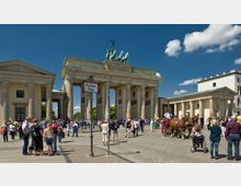 Das Bild zeigt das Brandenburger Tor in Berlin, ein neoklassizistisches Monument mit sechs Säulen und einer Quadriga auf der Spitze. Im Vordergrund befinden sich viele Menschen auf dem Pariser Platz sowie eine Pferdekutsche, die an einer sonnigen Tageskulisse vorbeifährt.