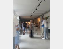 Unschärfes Bild einer modernen, hellen Innenraumkulisse mit Menschen, die sich bewegen. Im Hintergrund befindet sich eine Theke, und an der Wand rechts sind bunte Luftballons angebracht.