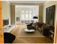 Wohnzimmer mit hellem Holzboden, einer großen braunen Couch, einem runden Couchtisch und einem Teppich. An der Wand hängt ein großes abstraktes Gemälde, links steht ein Flachbildfernseher auf einem niedrigen weißen Schrank, und im Hintergrund sind große Fenster mit Blick ins Grüne.