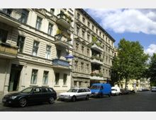 Straßenszene mit mehrstöckigen Altbauwohnhäusern im Gründerzeitstil, verziert mit Balkonen und Stuckelementen; parkende Autos stehen entlang des Kopfsteinpflasters, und Bäume säumen die Straße unter einem teilweise bewölkten Himmel.