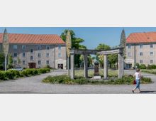 Eine parkähnliche Platzanlage mit einem steinernen Rundbogen und einer Büste auf einem Sockel in der Mitte. Im Hintergrund sind zwei gegenüberliegende Gebäude mit roten Ziegeldächern und schlichter Putzfassade zu sehen; umgeben von Bäumen und Büschen.