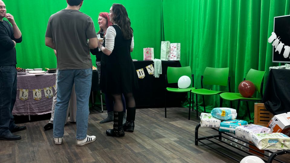 Indoor studio space with green-screen fabric covering the walls and bright fluorescent strip lights and acoustic foam panels on the ceiling. The room has wood-look flooring, a few chairs and tables with party decorations, balloons, and packaged supplies.