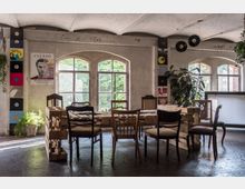 Ein rustikaler Raum mit großen Fenstern, die viel natürliches Licht hereinlassen. Der Raum ist mit unterschiedlichen Stühlen und einem Tisch aus Holzpaletten ausgestattet; an den Wänden befinden sich Plakate und Schallplatten, Pflanzen ergänzen das Interieur.