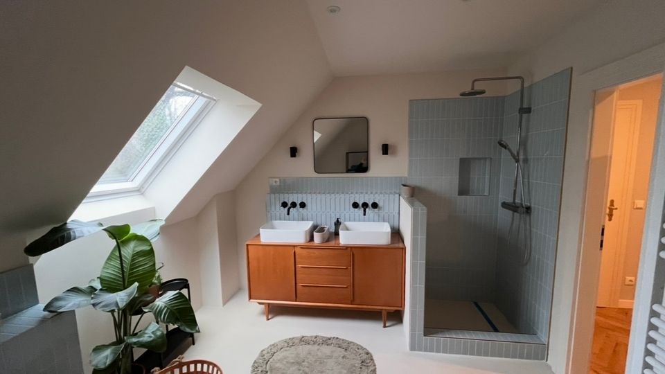 Modernes Badezimmer mit Dachschräge und großem Dachfenster. Der Raum verfügt über ein Doppelwaschbecken auf einem Holzunterschrank, eine offene Dusche mit blauen Wandfliesen, eine grüne Zimmerpflanze und helle Wände.