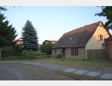 Ein kleines, einfaches Haus mit braunem Ziegeldach und gelblicher Fassade steht an einer gepflasterten Straße. Im Vorgarten befindet sich ein Gartenzaun, einige Pflanzen und ein großer Nadelbaum, umgeben von weiteren Bäumen und Gebäuden im Hintergrund.