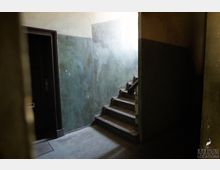 Treppenhaus in einem alten Gebäude mit einer dunklen Holztür links, grünlich-gestrichenen Wänden und einer schmalen Treppe mit abgenutztem Holzgeländer, die nach oben führt. Wenig Beleuchtung durch ein oberes Fenster.