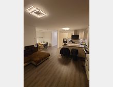 Modernes Wohnzimmer mit offener Küche und Essbereich. Der Raum hat einen dunklen Laminatboden, ein großes Sofa, eine Küchenzeile mit weißen Schränken und eine Esstischecke mit Hängelampen.