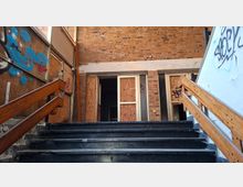 Außentreppe mit dunklen Stufen und Holzgeländern führt zu einem Eingang in einem Backsteingebäude. Fenster und Türen sind mit Sperrholzplatten vernagelt, die Wände zeigen Graffiti und teils beschädigte Oberflächen.
