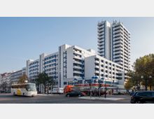 Mehrstöckiger Wohn- und Geschäftsgebäudekomplex in moderner Architektur mit weißen Fassaden und Balkonen, flankiert von einem hohen Wohnturm. Im Vordergrund sind eine Straße mit parkenden und fahrenden Fahrzeugen sowie ein Supermarkt sichtbar.