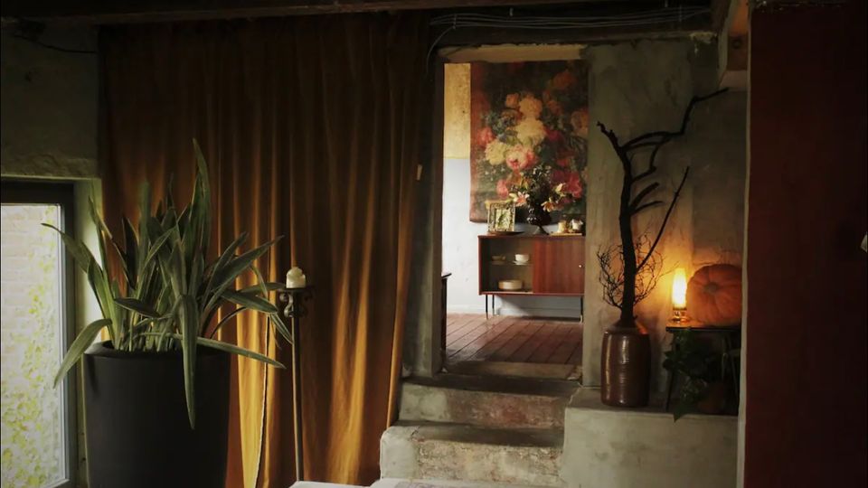 Raum mit rustikalem Interieur, bestehend aus einem großen Topfpflanzengefäß, goldfarbenem Vorhang und einer Treppe, die in einen Nebenraum mit Holzboden führt, wo ein Sideboard und ein großformatiges Blumenbild sichtbar sind. Ein leuchtender Lampenschirm sowie dekorative Elemente wie ein Kürbis und ein dürrer Ast in einer Vase ergänzen die Szenerie.