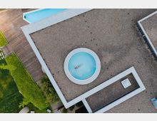 Draufsicht auf ein modernes Gebäude mit Kiesdach, in dessen Zentrum sich ein runder Swimmingpool befindet, in dem eine Person auf dem Rücken schwimmt. Umgeben ist das Gebäude von einem Holzdeck und gepflegten grünen Rasenflächen.