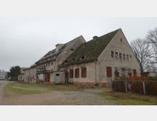 Ein verlassenes, zweigeschossiges Gebäude mit steilem, mit Moos bewachsenem Dach und teils zugenagelten Fenstern und Türen. Die Fassade ist stark verfallen, umgeben von unbefestigtem Boden, niedrigen Sträuchern und kahlen Bäumen im Hintergrund.