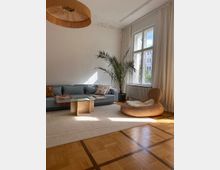 Wohnzimmer mit hellem Holzboden und hohen Decken, dekoriert mit einer großen, grauen Couch, einem Glastisch auf einem beigen Teppich, einem geflochtenen Sessel mit Fellauflage und einer grünen Zimmerpflanze neben großen Fenstern mit weißen Vorhängen.